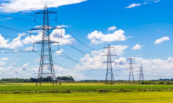 A row of tall electricity pylons with power lines crossing a lush green field under a bright blue sky with clouds
