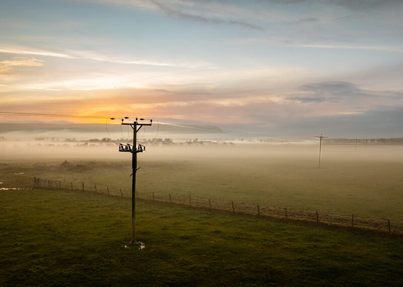 A wooden electricity pole with overhead power lines crossing a misty green field at sunrise