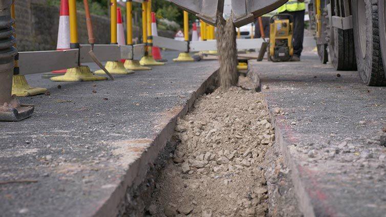 A lorry tipping gravel into a dug-up trench in a road during gas works, with traffic cones and barriers