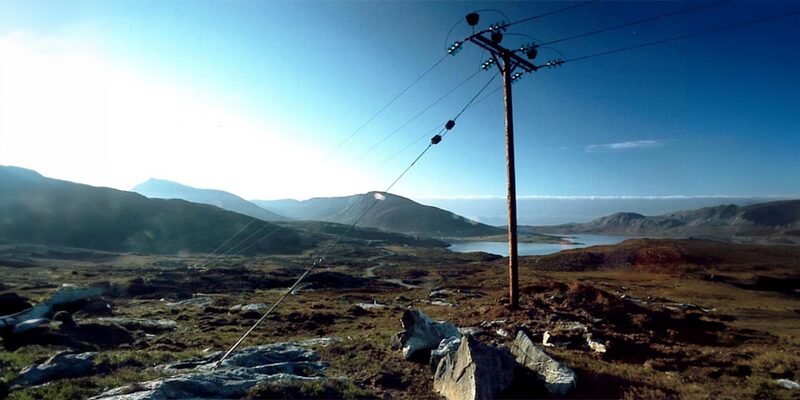A wooden electricity distribution pole with overhead power lines crossing a rocky moorland landscape with hills and a loch