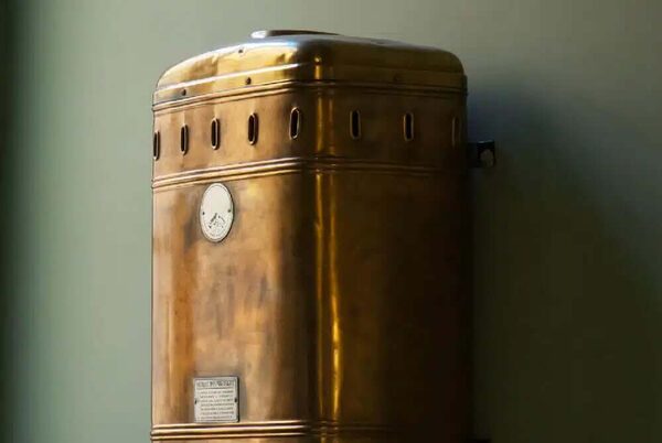 An old brass-cased wall-mounted gas water heater or boiler against a green wall