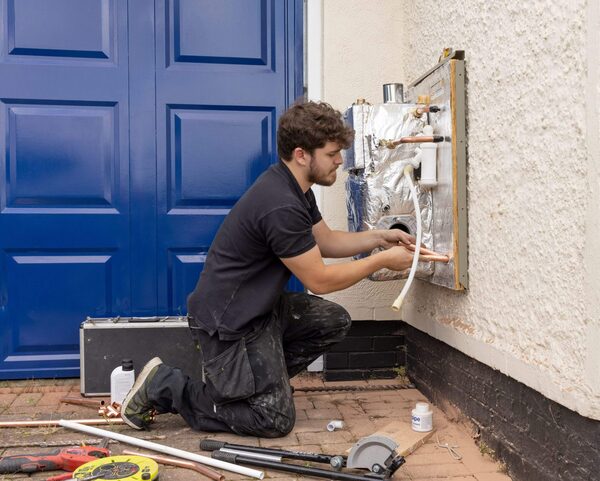 A heating engineer kneeling outside a house installing copper pipework for a wall-mounted boiler unit