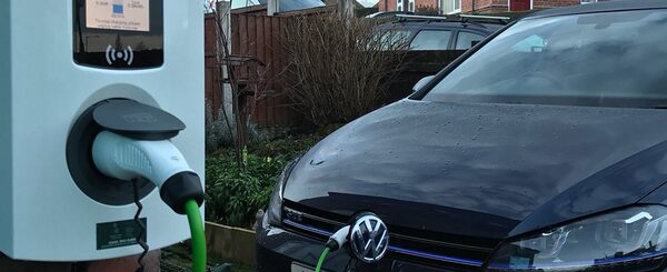 A dark Volkswagen car plugged in with a green cable to a white home EV charge point on a driveway