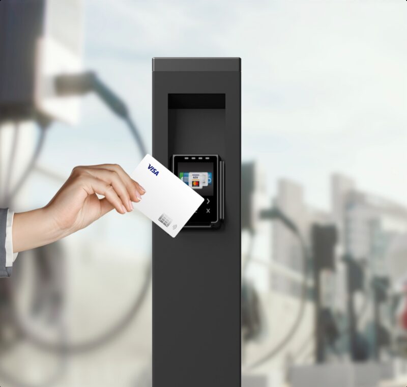 A hand tapping a Visa contactless card on the payment terminal of a black public EV charge point pedestal