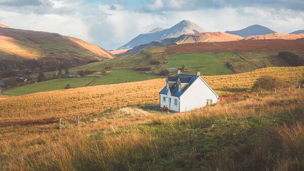 A white croft house with a blue roof standing alone in a grassy glen with mountains behind, in the Scottish Highlands