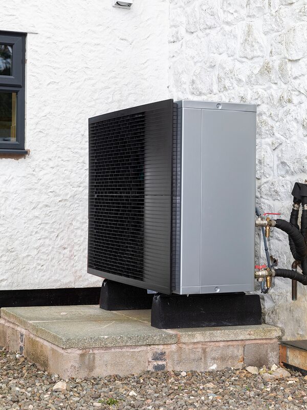 An air source heat pump unit installed outside a white stone house on a paved base
