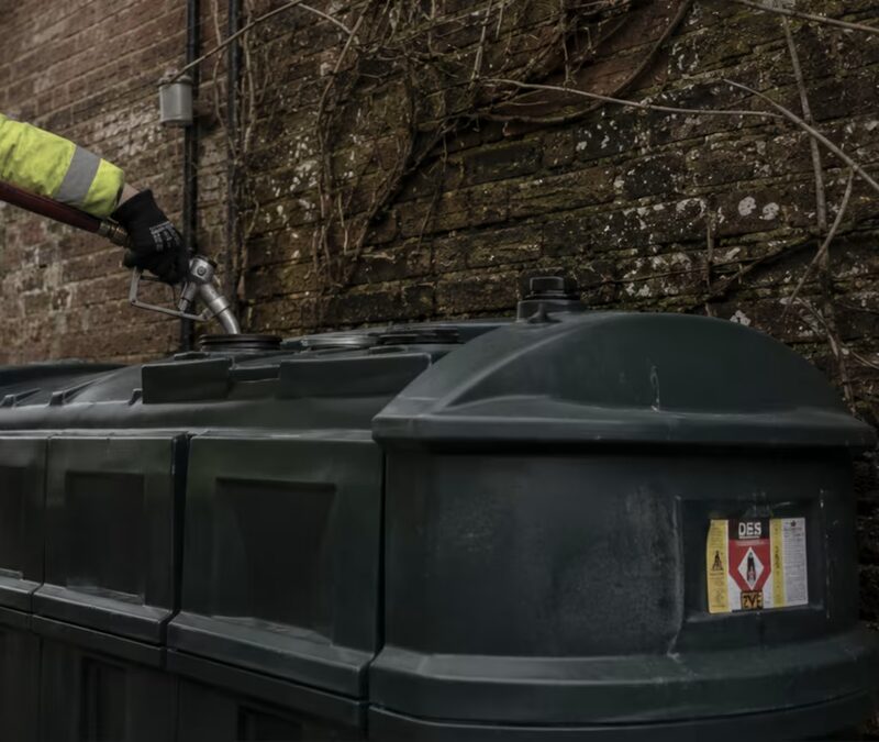 A gloved hand filling a green domestic heating oil tank from a delivery hose against a brick wall
