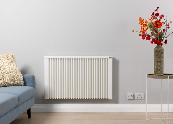 A white electric panel heater mounted on a grey living room wall beside a blue sofa and a vase of flowers