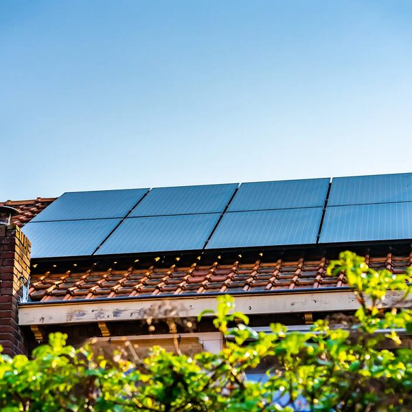 Solar panels installed on the tiled roof of a brick house, seen from below with greenery in the foreground