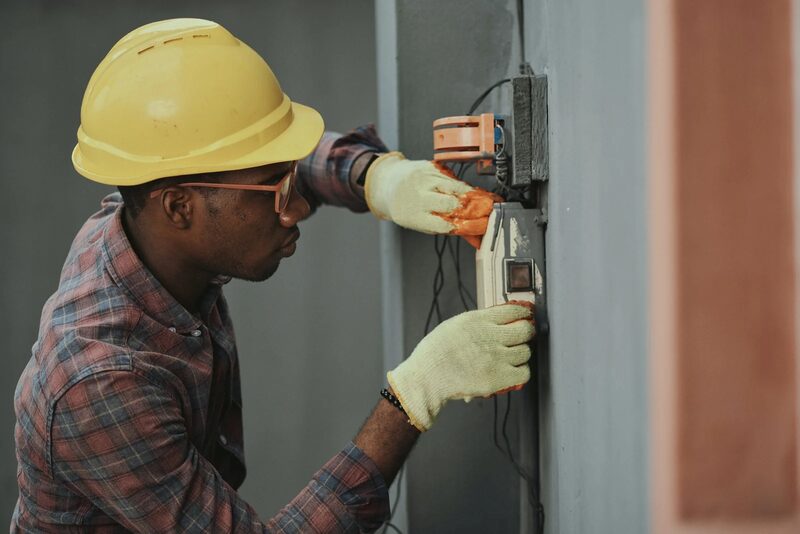 An electrician in a yellow hard hat and gloves working on an electrical meter or switch box mounted on a wall