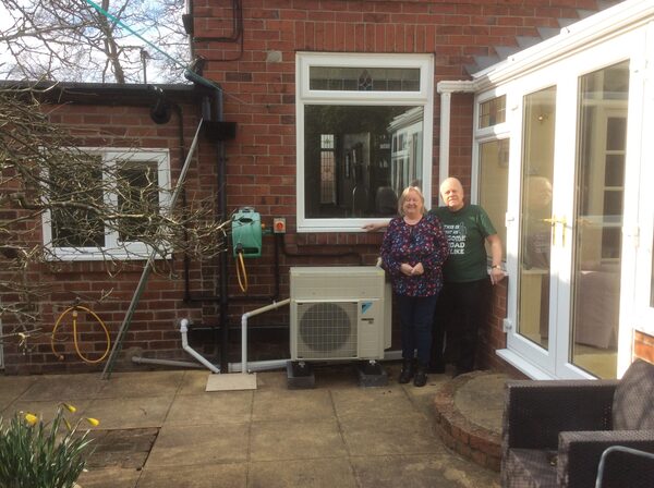An older couple standing beside a Daikin air source heat pump unit installed outside their brick house