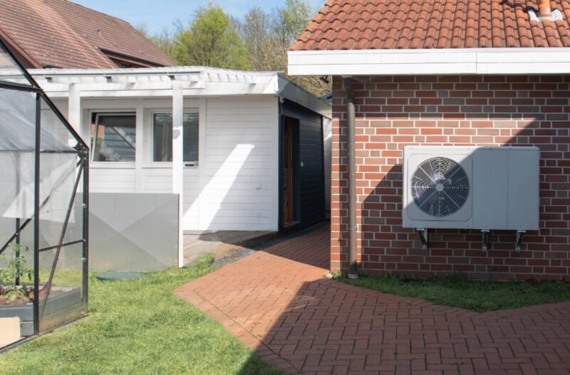 An air source heat pump unit mounted on the brick wall of a house next to a greenhouse and garden path