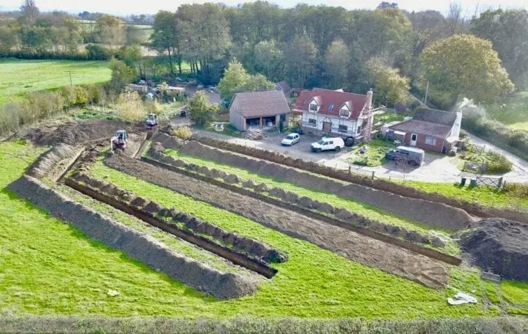 Aerial view of trenches being dug in a field beside a rural house for a ground source heat pump ground array installation
