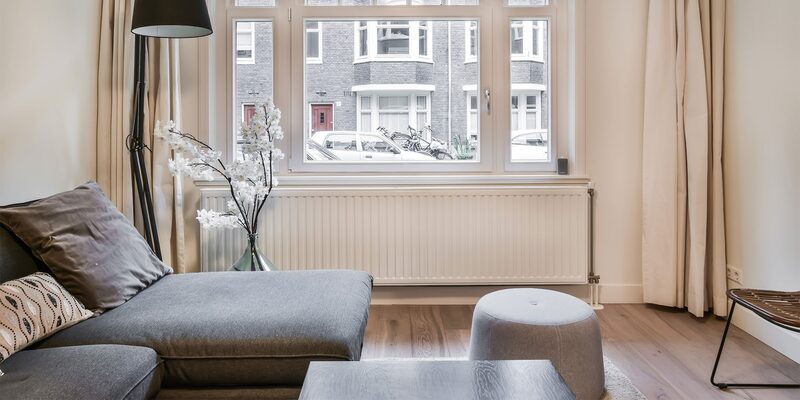 A white panel radiator under a window in a living room with a grey sofa and curtains