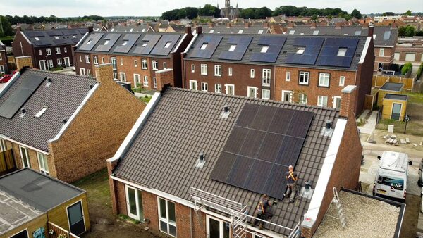 Installers fitting solar panels on the tiled roof of a brick house in a residential neighbourhood with many solar-covered roofs