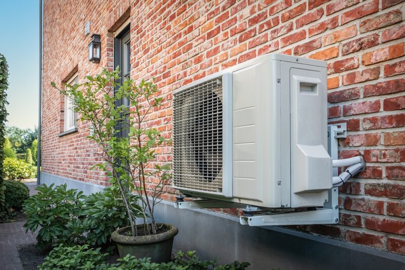 An air source heat pump unit mounted on the exterior brick wall of a house beside a potted shrub