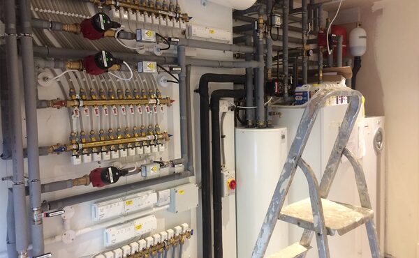 A plant room with an installed ground source heat pump system, including manifolds, pumps, pipework, a hot water cylinder and a step ladder