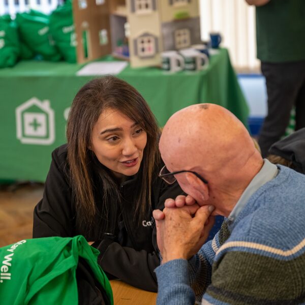 A woman adviser talking with an elderly man at a Cadent Foundation fuel poverty support event with green branded bags and a model house on a table