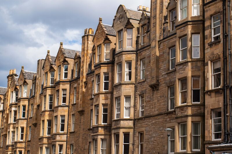 A row of traditional Scottish sandstone tenement buildings with bay windows and slate roofs under a cloudy sky