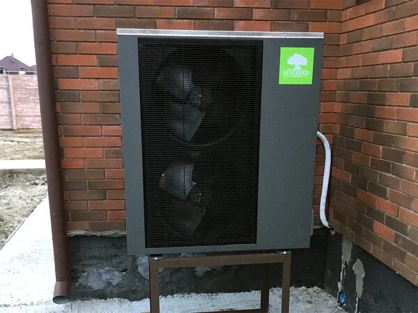 A Mycond air source heat pump unit installed on a metal stand outside a brick house wall