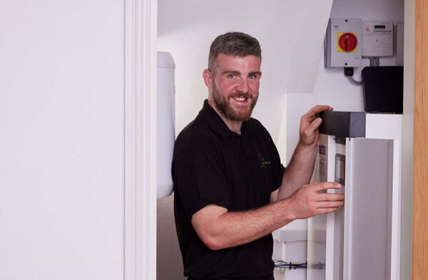 A smiling engineer in a black polo shirt installing an indoor heat pump or hot water cylinder unit in a UK home airing cupboard