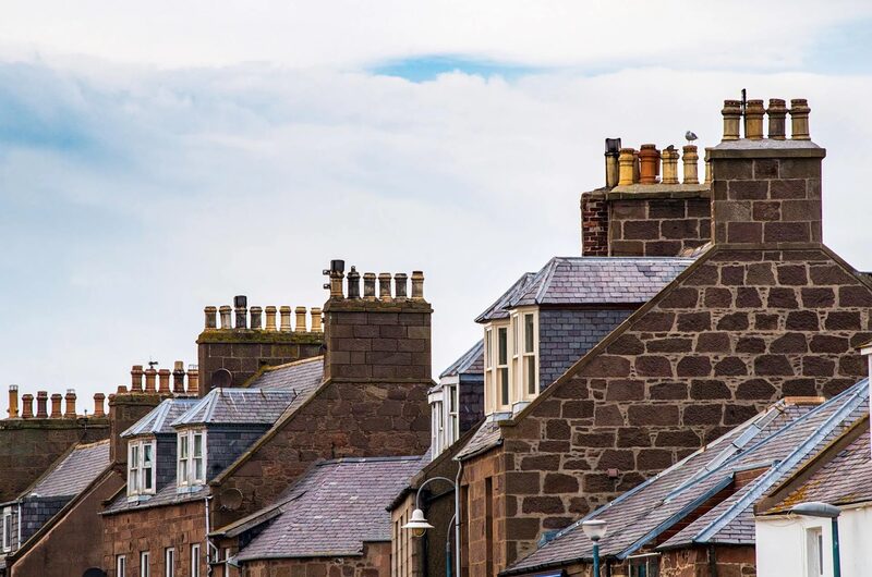 A row of traditional stone-built terraced houses with slate roofs and multiple brick chimney stacks under a cloudy sky
