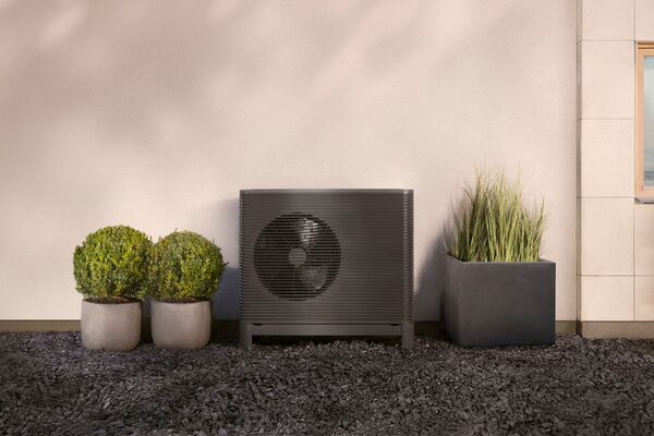 A dark grey air source heat pump unit installed outside a house wall, flanked by potted shrubs and grasses on a gravel bed