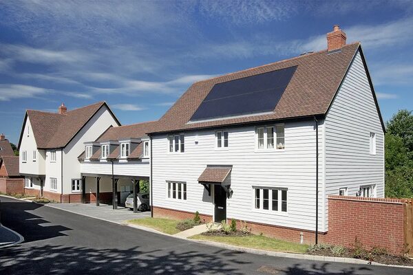 A white weatherboarded UK house with solar panels on its tiled roof, next to a carport and neighbouring homes