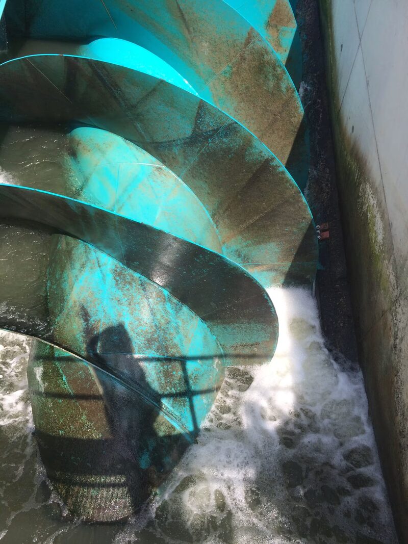A large turquoise Archimedes screw hydropower turbine turning in churning water beside a concrete wall