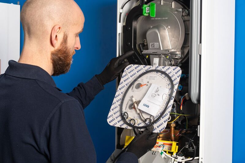 A technician in gloves servicing the inside of a Baxi boiler, holding the front panel with a Baxi-branded gasket and instruction sheet