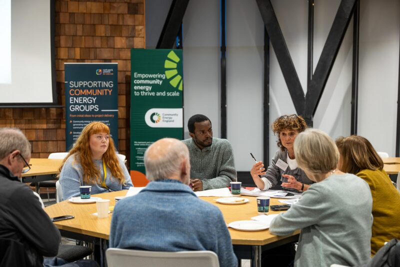 People in discussion around a table at a community energy event with Community Energy England banners behind them