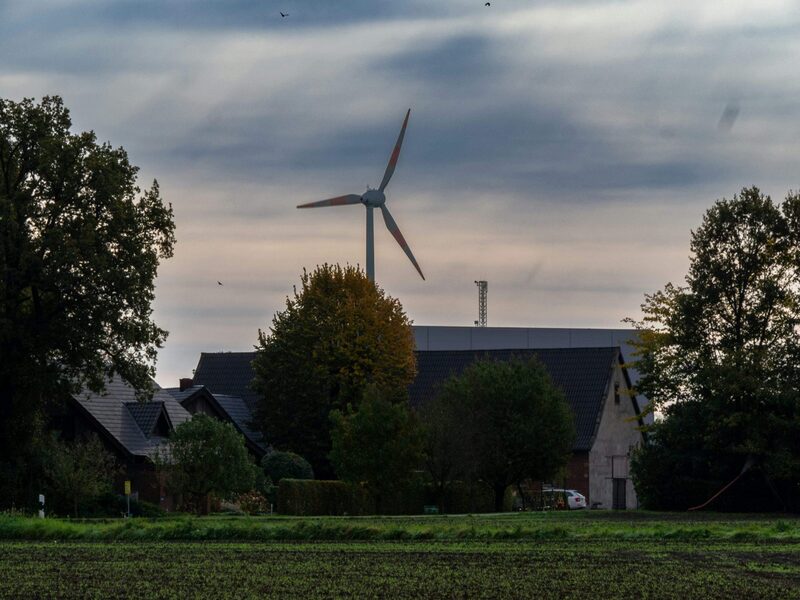A wind turbine rising above village rooftops and trees behind a field under an overcast sky