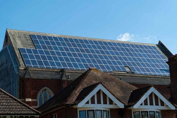 A large array of solar panels installed on the pitched roof of a brick community building, seen above neighbouring tiled house roofs under a blue sky