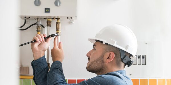 A technician in a white hard hat using a spanner to connect pipework on a wall-mounted gas boiler in a kitchen
