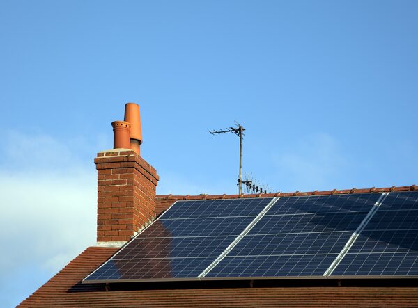 Solar panels installed on a tiled house roof beside a brick chimney and TV aerial under a blue sky