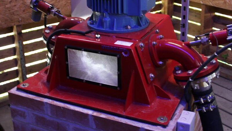 A red micro-hydro turbine generator with blue motor mounted on a brick plinth inside a wooden turbine house