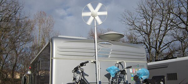 A small white wind turbine mounted on a pole above a silver caravan with bicycles on the rear rack