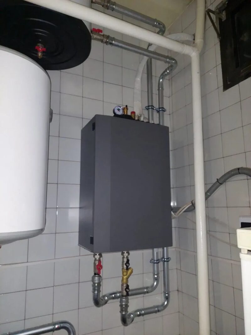 A grey wall-mounted heating manifold/hydro box installed on a tiled utility wall next to a white hot water cylinder, with copper and plastic pipework