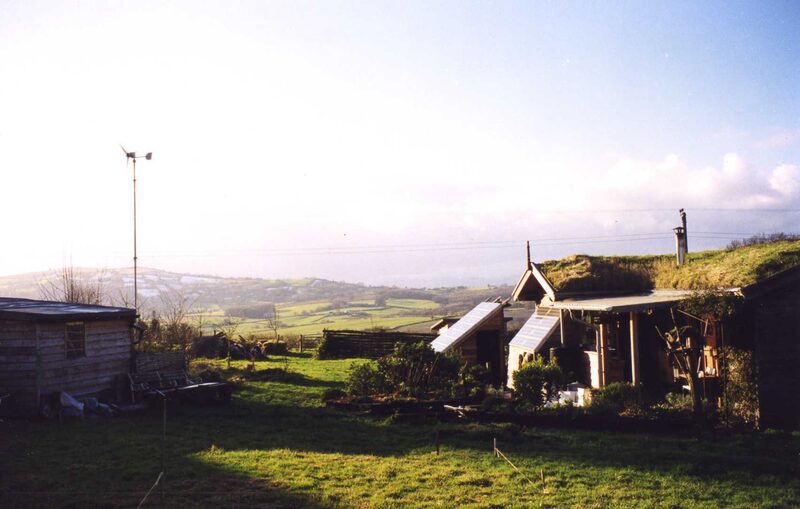 An off-grid rural homestead with a small wind turbine on a pole and solar panels beside turf-roofed buildings overlooking countryside