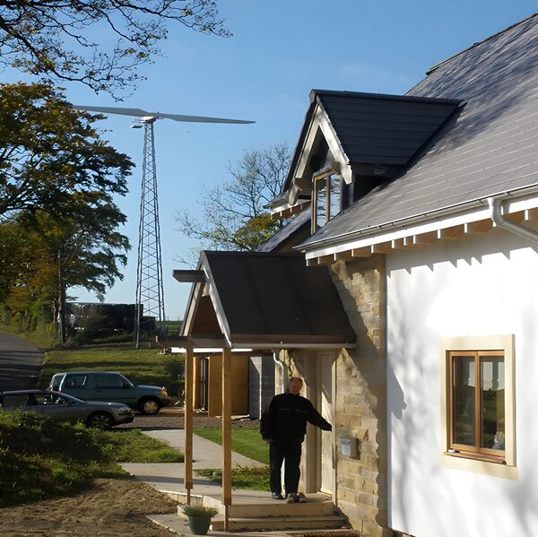 A man at the door of a modern eco-house with a small wind turbine on a lattice tower behind it
