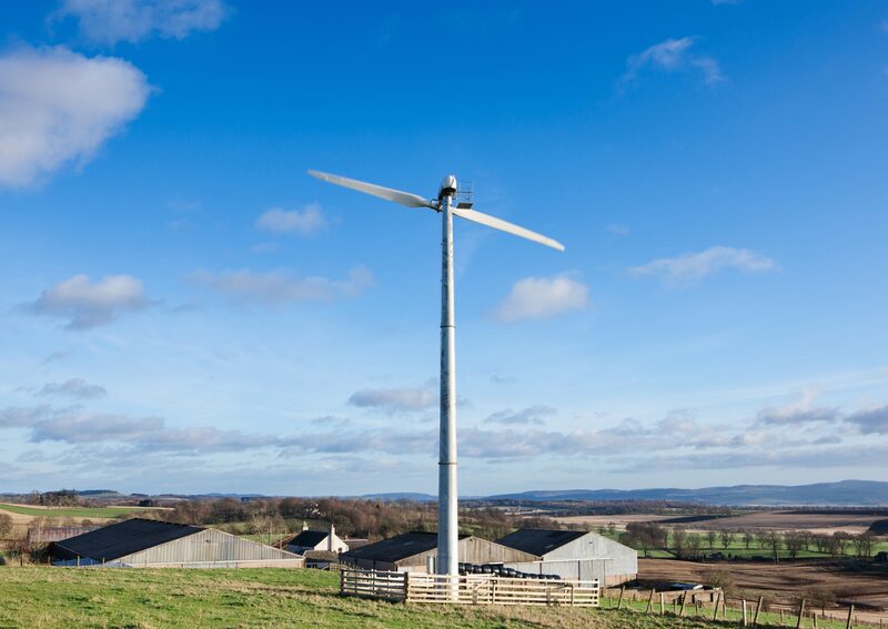 A small wind turbine on a tall tower in a rural farm landscape with barns and fields under a blue sky