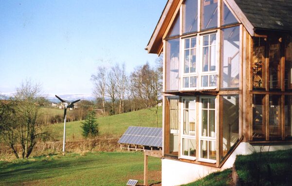 A timber-framed glass-fronted eco house in countryside with a small wind turbine and ground-mounted solar panels beside it