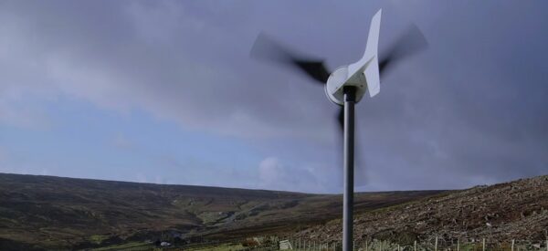 A small white wind turbine spinning on a pole in a hilly rural landscape under a cloudy sky