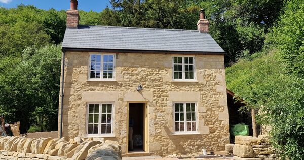A stone Grade II listed cottage with a slate roof, surrounded by trees and a dry stone wall