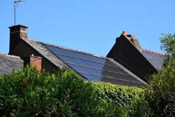 Black solar panels fitted on the slate roof of a house, seen above trees under a blue sky
