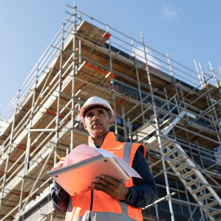 A building inspector in a hard hat and hi-vis vest holding a clipboard in front of a scaffolding-covered building under construction