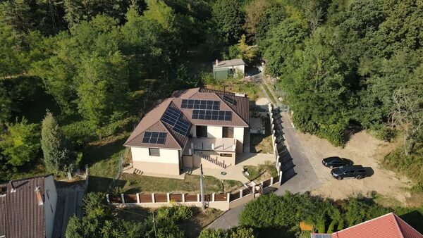An aerial view of a detached house with solar panels on its tiled roof, surrounded by trees and a fenced garden