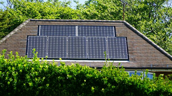Solar panels installed on the tiled roof of a house, partly hidden behind green hedges and trees