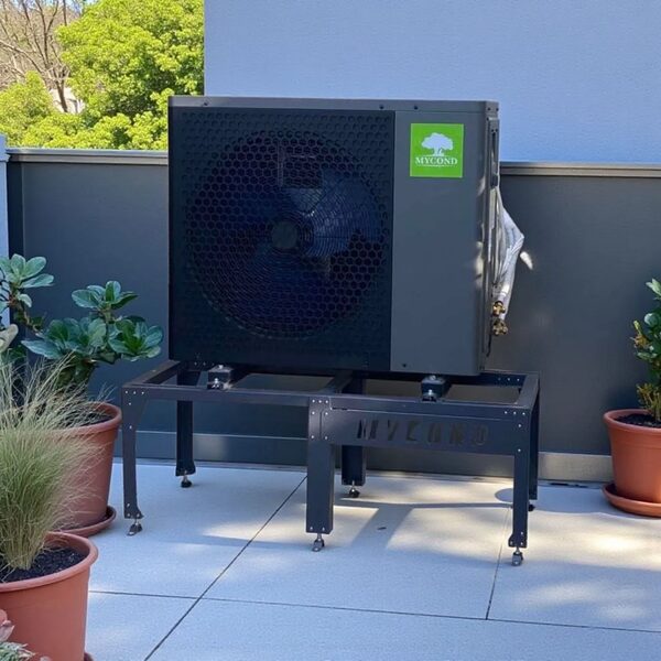 A Mycond split air-source heat pump outdoor unit installed on a black mounting frame on a paved terrace beside a house wall, with potted plants around it.
