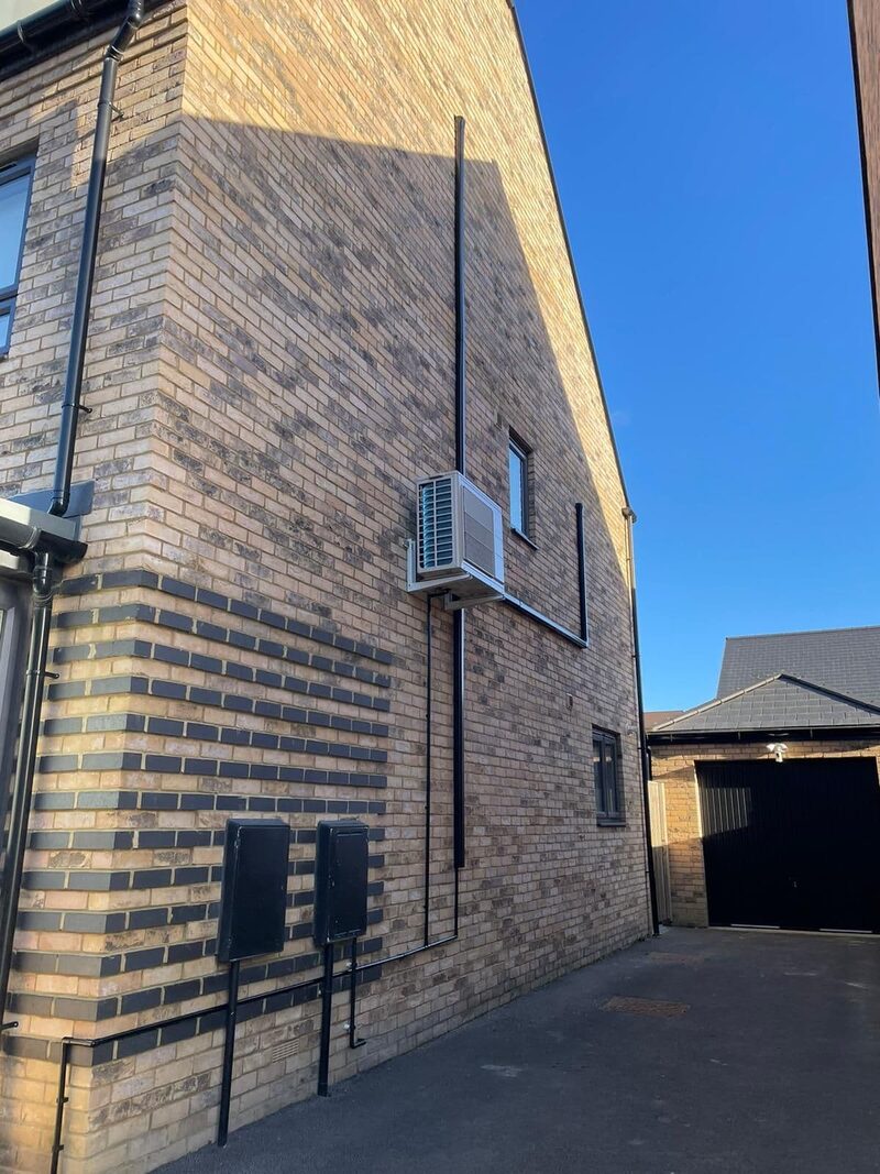 An outdoor air conditioning condenser unit mounted high on the brick side wall of a modern UK house